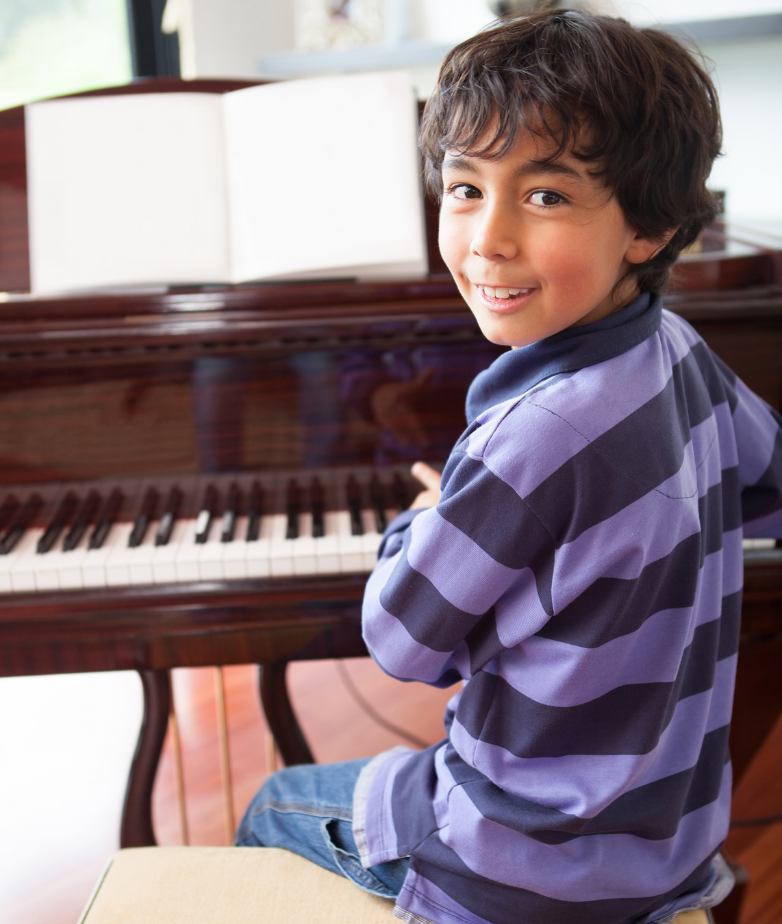 A young boy smiling while playing the piano during a music lesson. He is wearing a purple striped shirt and sitting at a grand piano with an open sheet of music in front of him, in a bright and welcoming space.