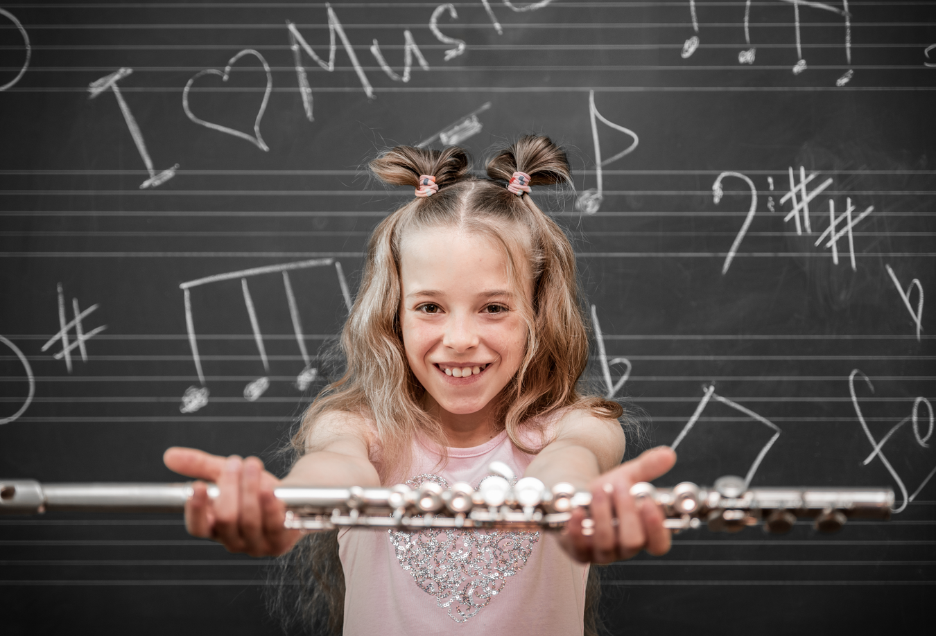 A smiling girl holding a silver flute in front of a blackboard filled with musical notes and the phrase 'I ❤️ Music.' This image highlights the joy of learning music and represents the availability of rental beginning band instruments.